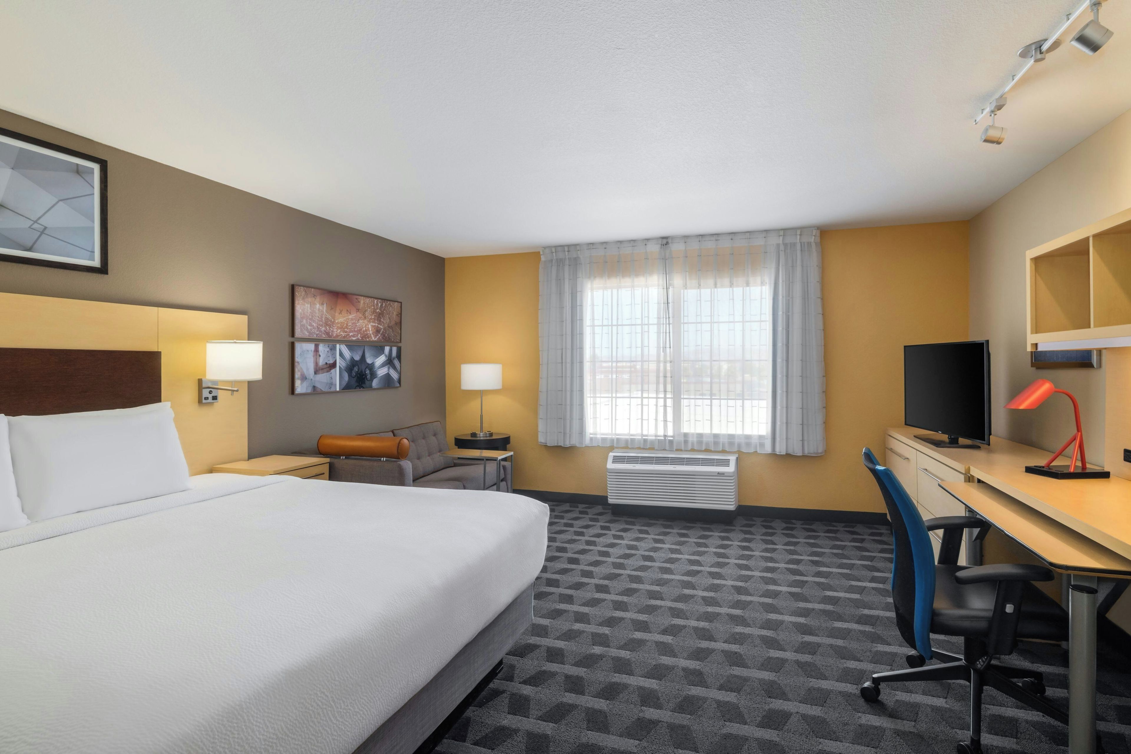 A hotel room with a bed, desk, chair, TV, and artwork on the walls.