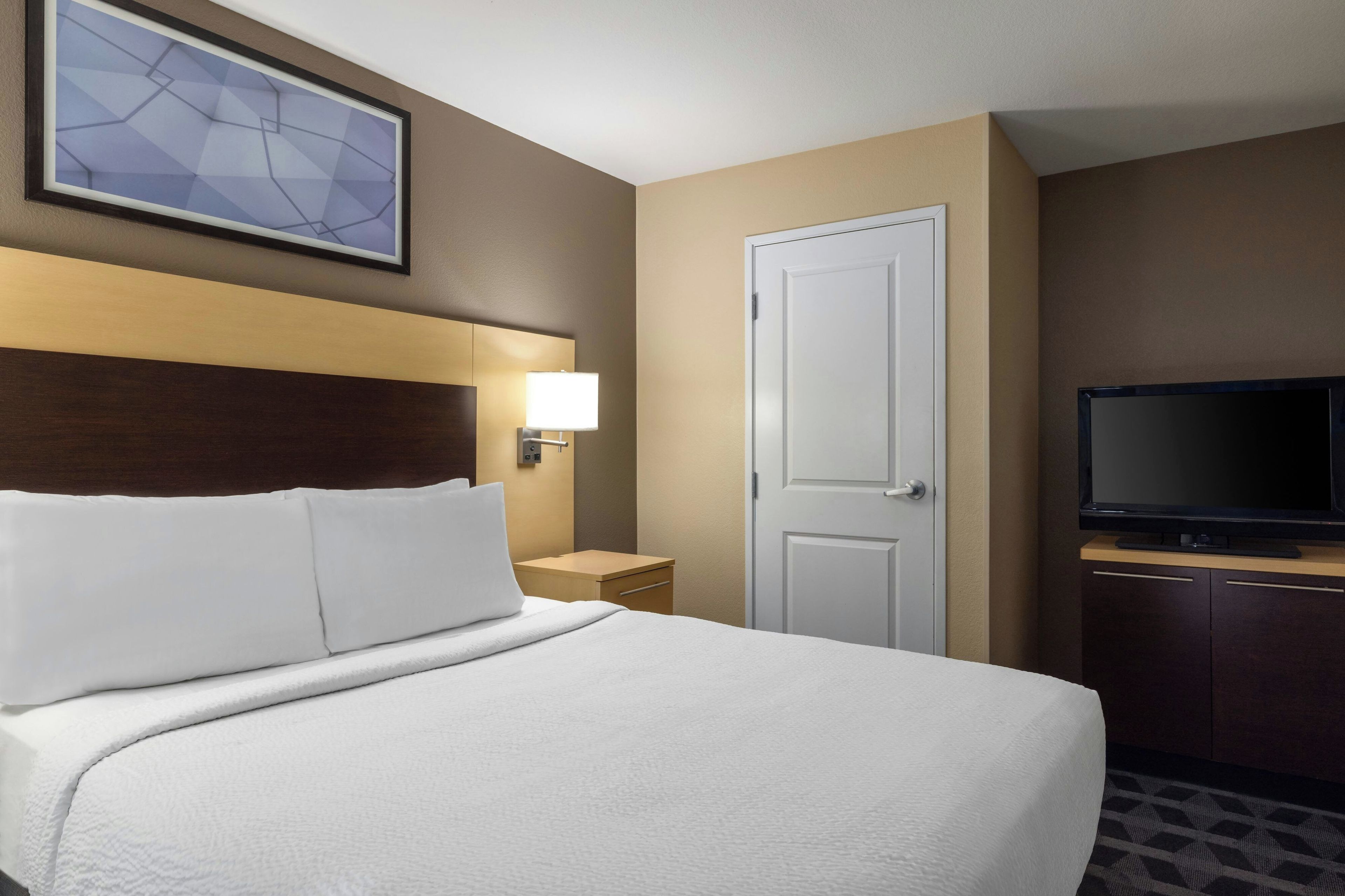 A hotel room with a bed, a TV, a door, and a wall-mounted light.