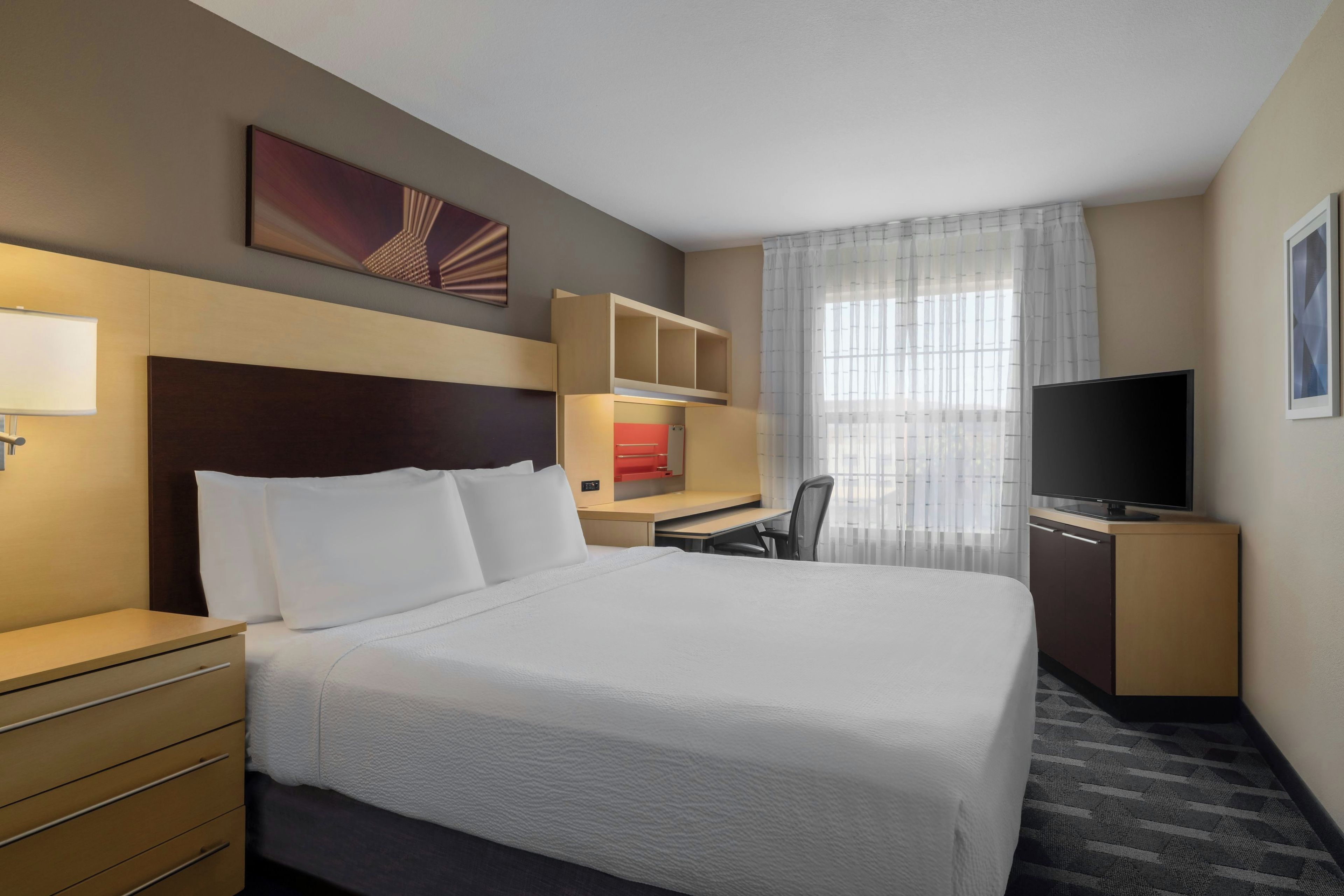 A hotel room with a large bed, a desk with a chair, a television, and a window with sheer curtains.