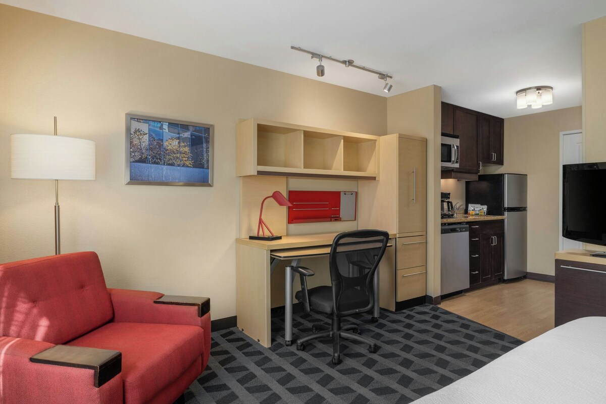 Habitación de hotel con cama, escritorio y silla, sofá rojo, zona de cocina con microondas y refrigerador, y un cuadro en la pared.