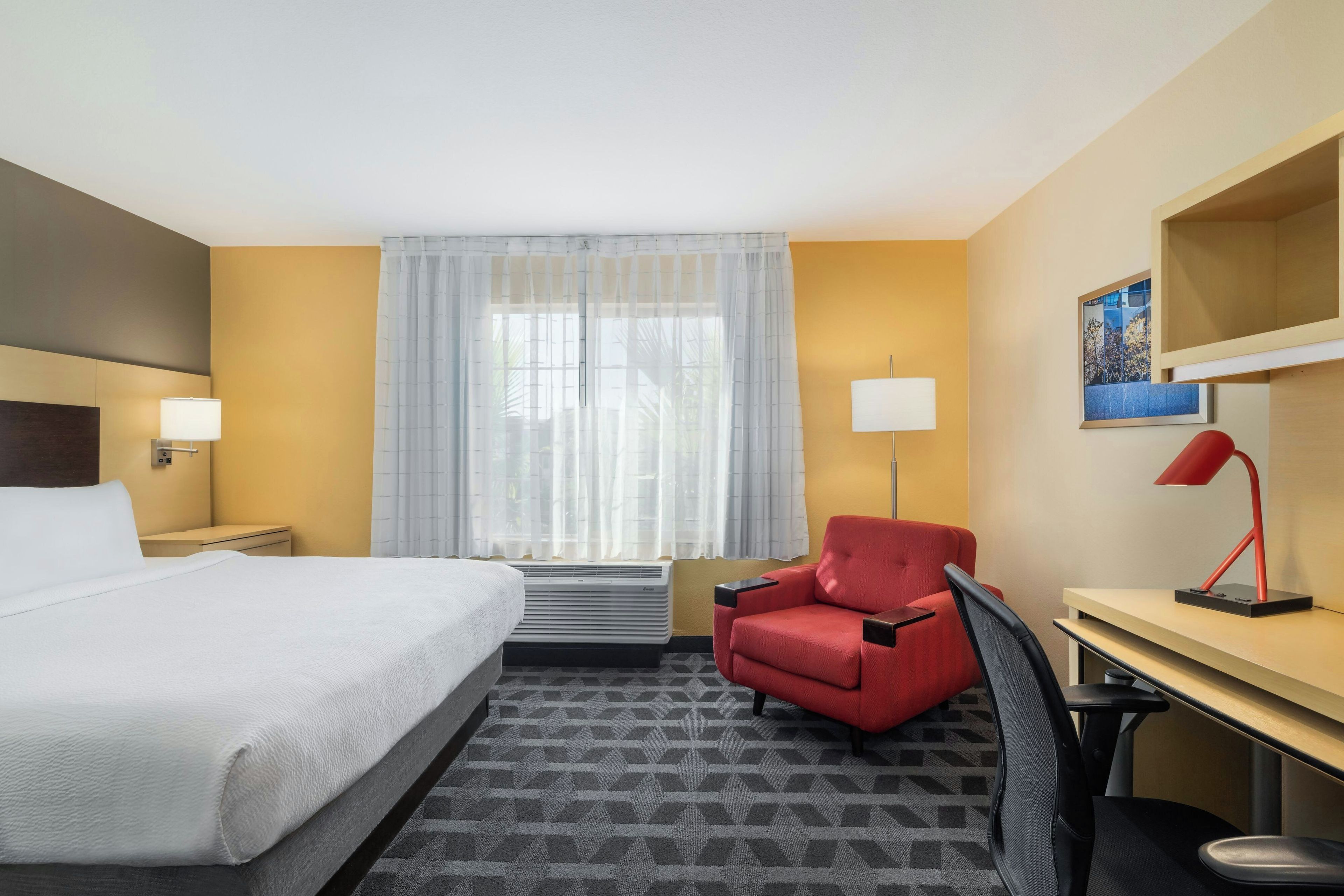 A hotel room with a bed, a desk, a chair, a lamp, a window with sheer curtains, and a wall-mounted picture.
