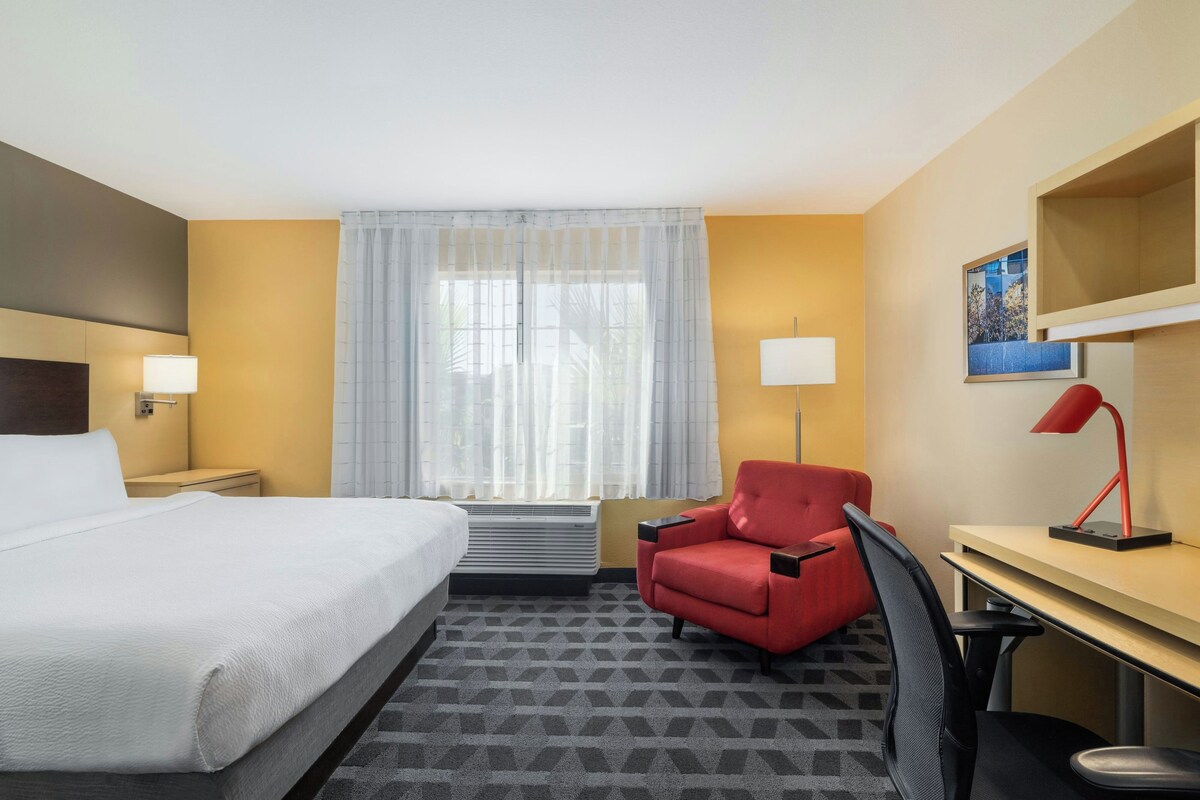 A hotel room with a bed, a desk, a chair, a lamp, a window with sheer curtains, and a wall-mounted picture.