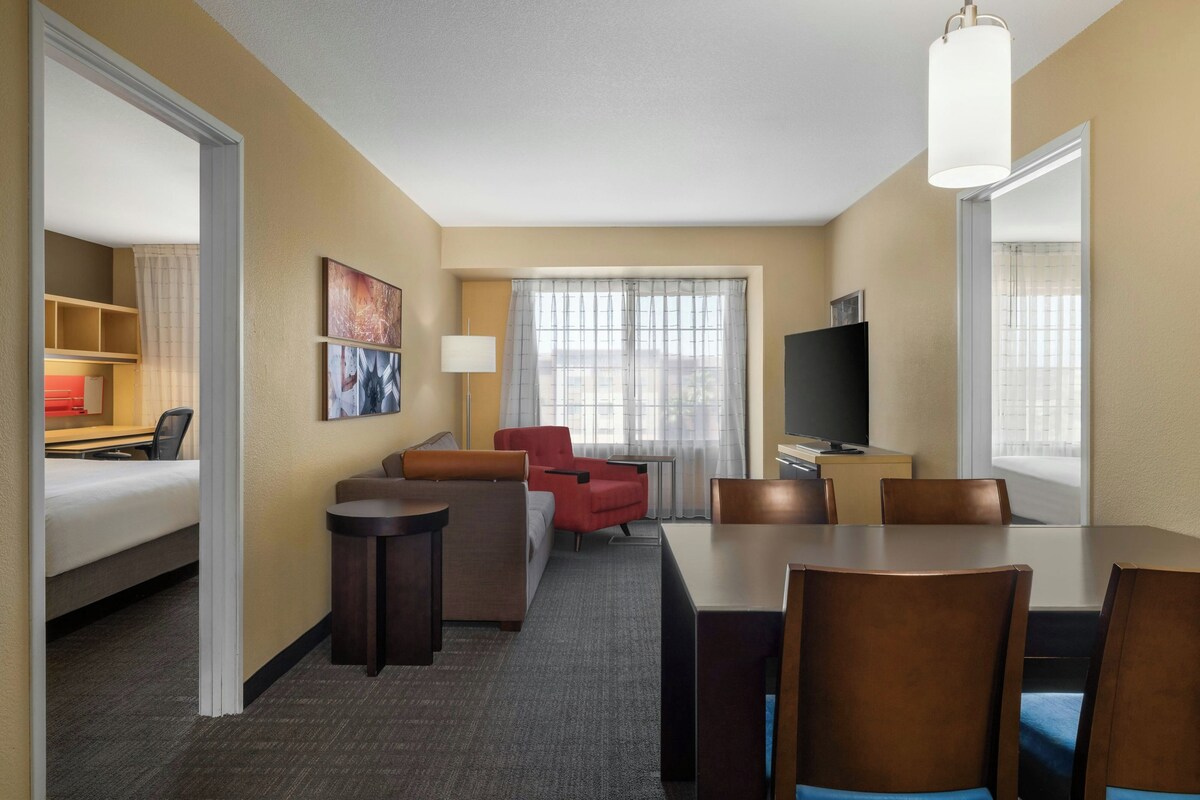 A hotel room with a bed, a desk, a TV, a dining table with chairs, and a window with curtains.