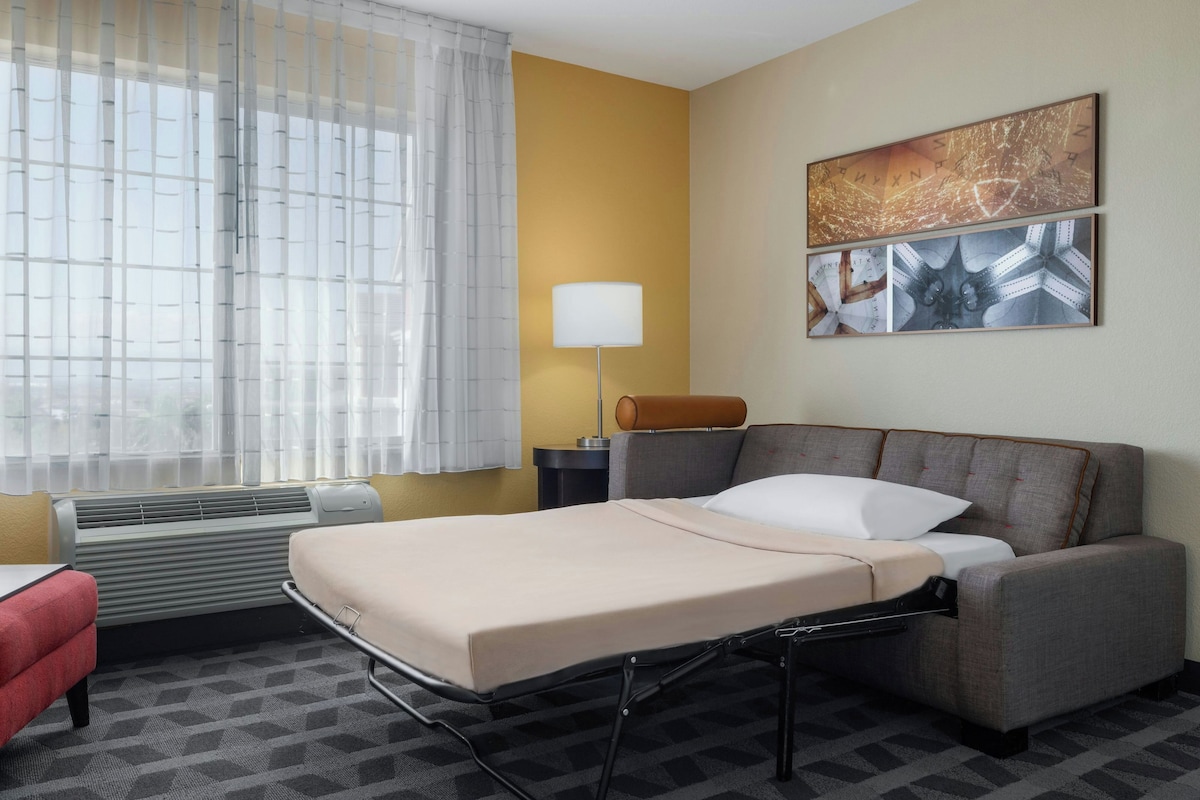 A hotel room with a bed, a desk, a chair, a lamp, and artwork on the wall.