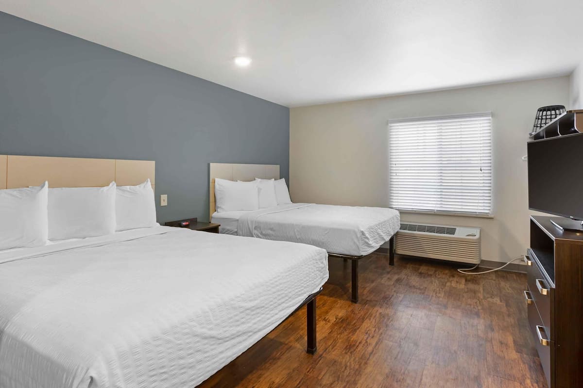 A hotel room with two beds, a wooden floor, a window with blinds, a heater, and a TV on a stand.
