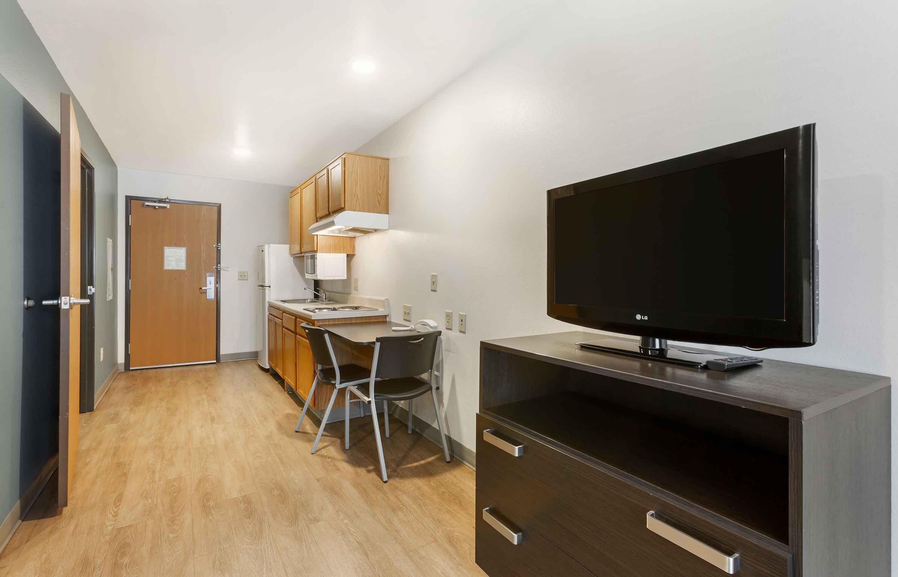 A compact hotel room with a kitchenette, a flat-screen TV, a desk with a chair, and a wardrobe.