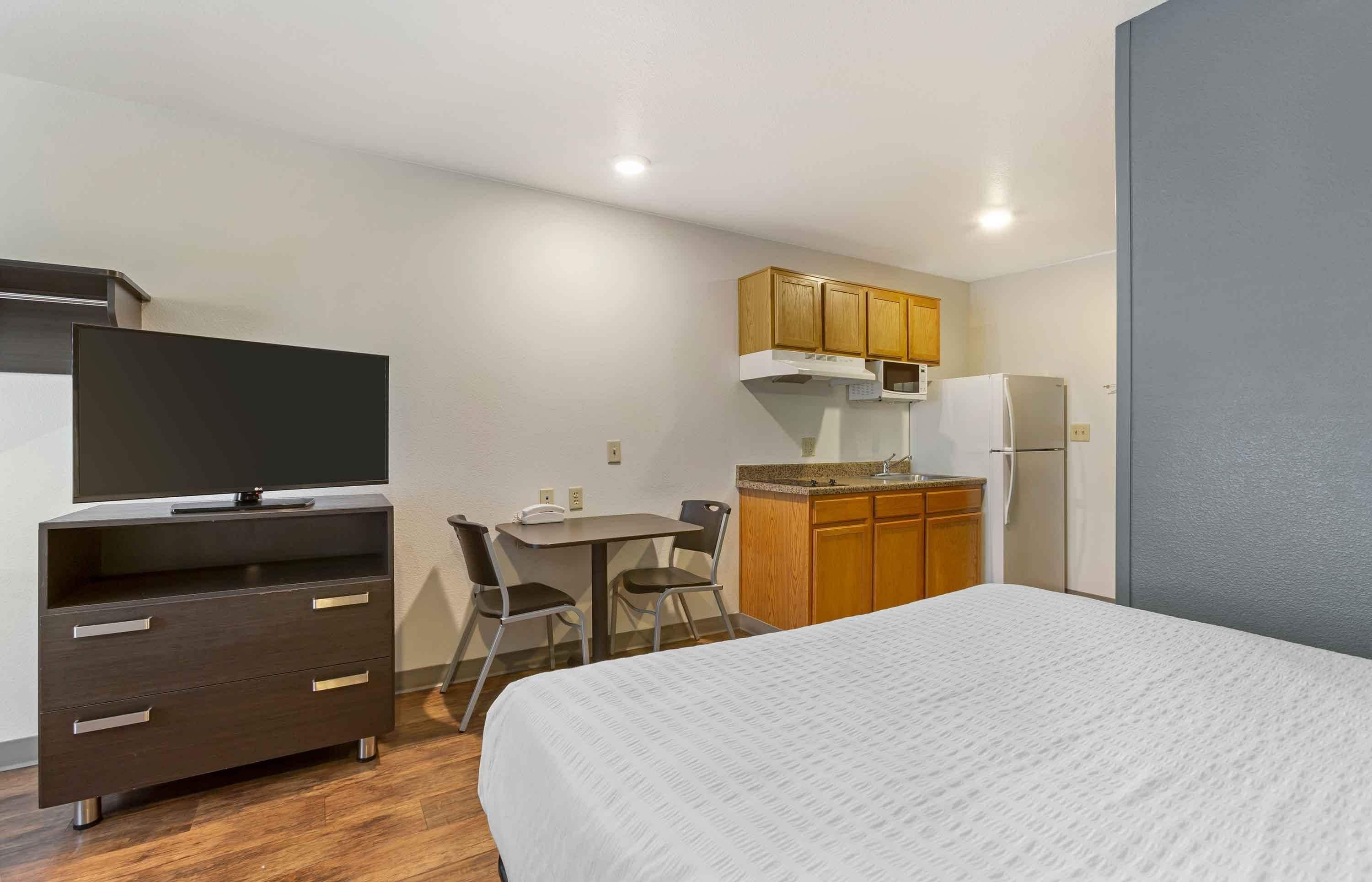 Una habitación compacta con una cama, un televisor sobre un tocador, una kitchenette pequeña con microondas y refrigerador, y un rincón comedor con mesa y sillas.