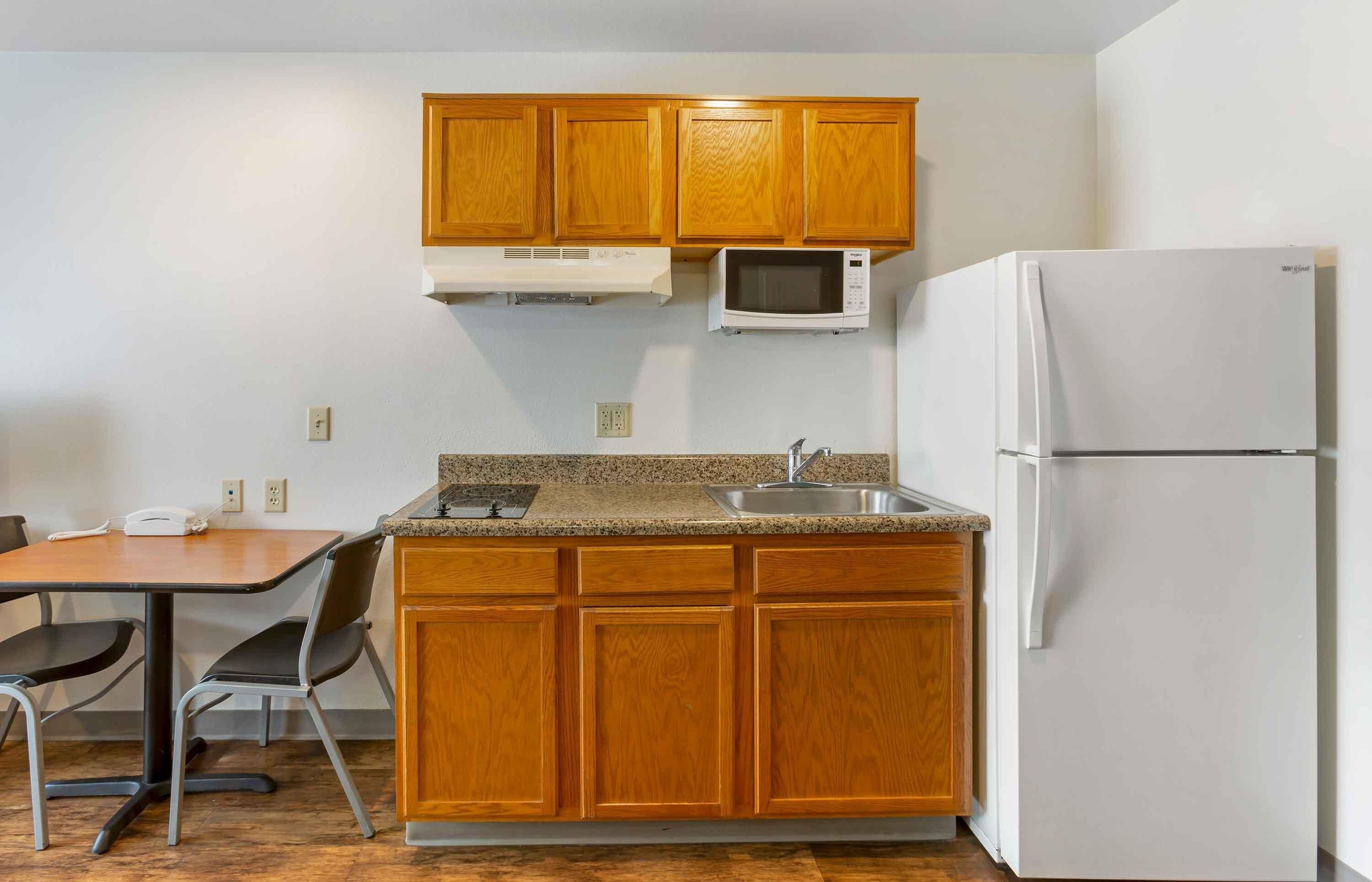 Una cocina compacta con armarios de madera, microondas, refrigerador, encimera de granito, lavamanos y una mesa de comedor con sillas.