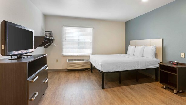A modern hotel room with a bed, a TV on a dresser, a heater, and a window with blinds. - Extended Stay America Select Suites - Phoenix - North (Phoenix)