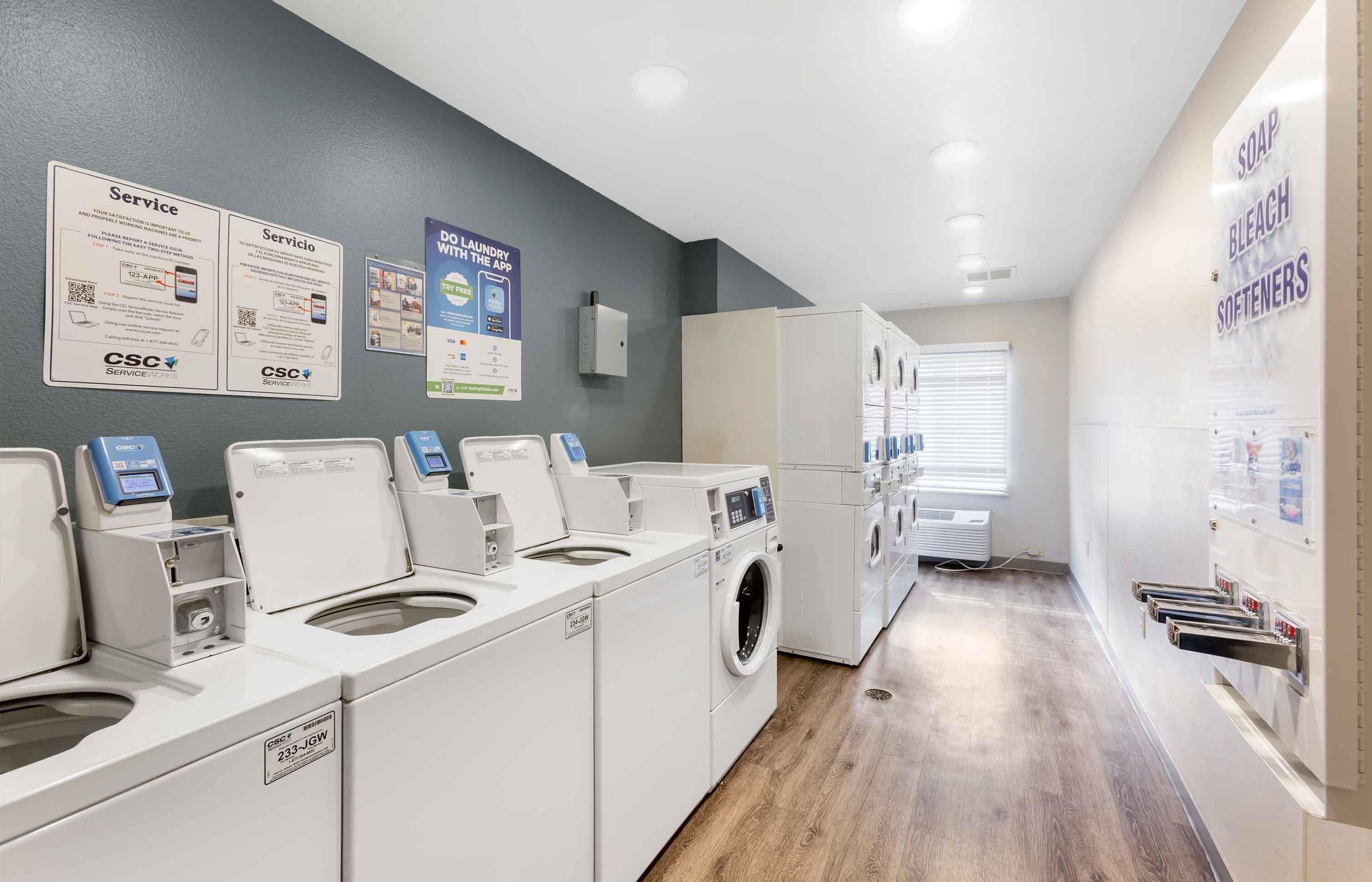 A laundry facility with multiple washing machines and dryers, a service sign, and a wall-mounted information board.