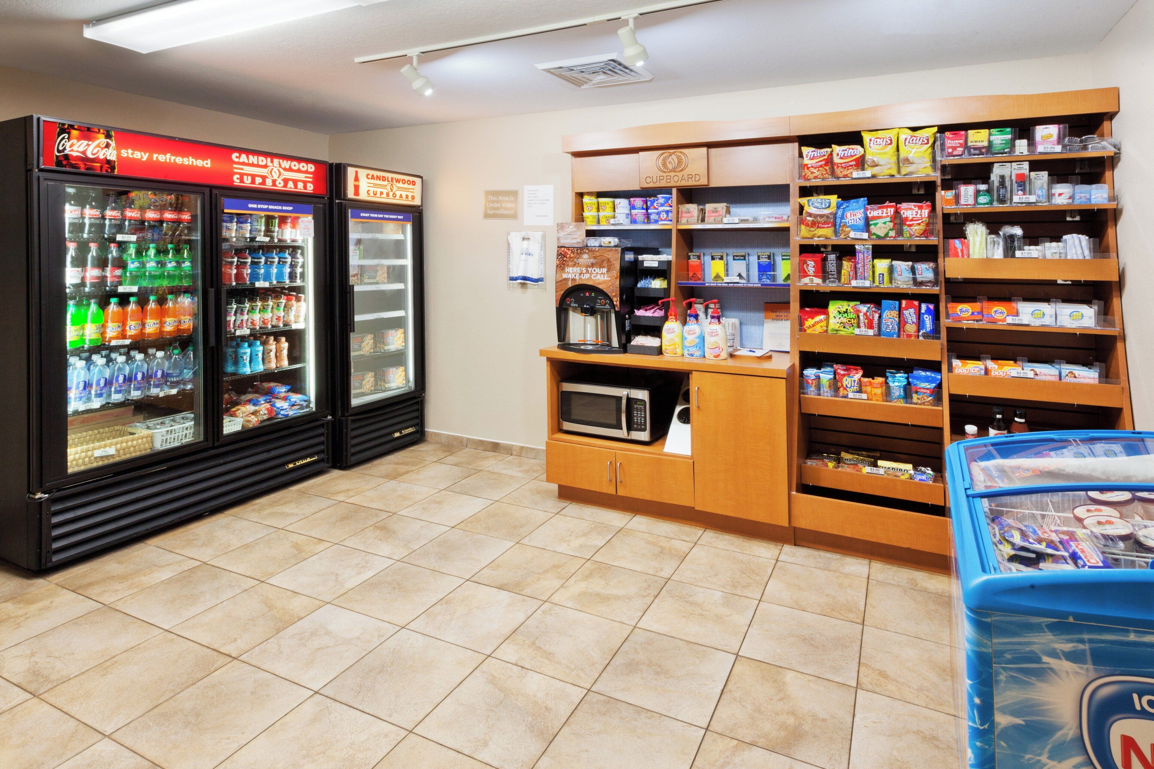 A snack and beverage vending area with Coca-Cola and Candlewood Cupboard machines, a microwave, and shelves stocked with snacks and beverages.