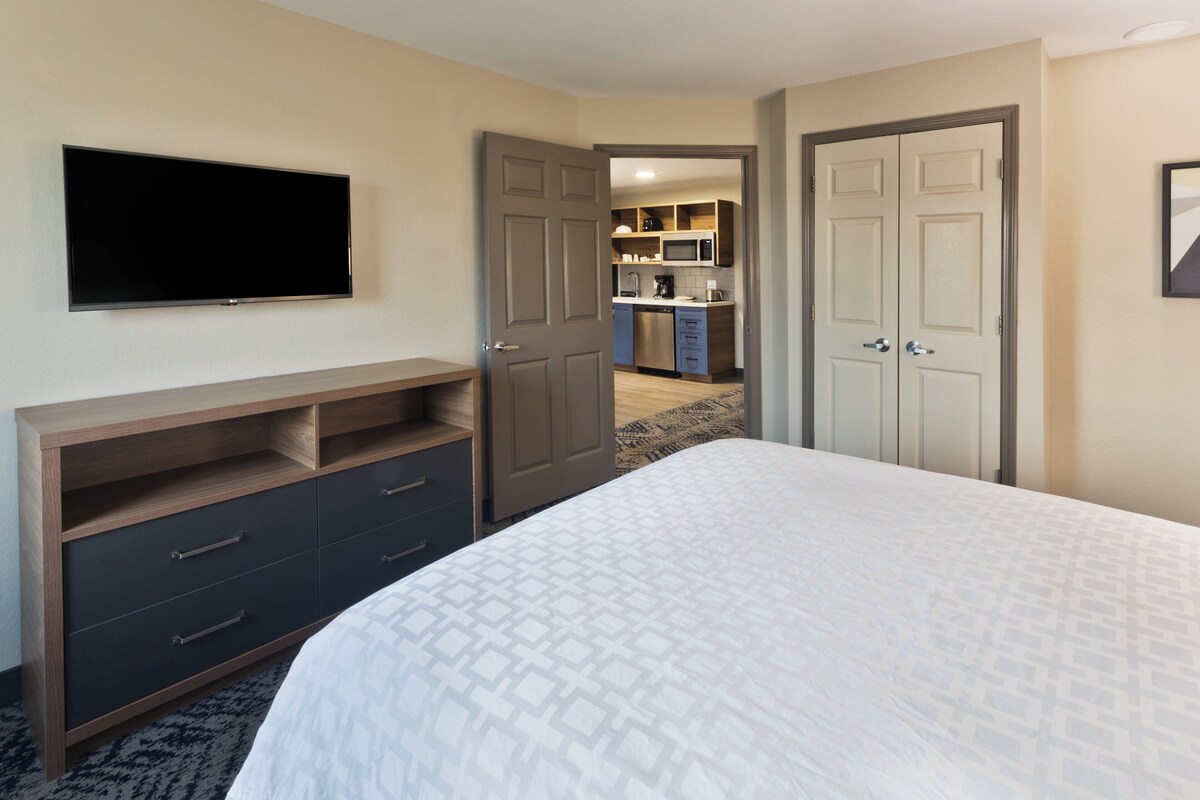 Habitación de hotel con una cama, un televisor montado en la pared, un aparador de madera y una cocina visible a través de una puerta abierta.