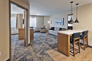 Een hotelkamer met een kitchenette, een bank, een eethoek met barstoelen, een kamerbreed tapijt en een groot raam met gordijnen.