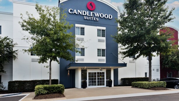 A modern hotel building with a prominent "CANDLEWOOD SUITES" sign, featuring a curved entrance and a glass door.