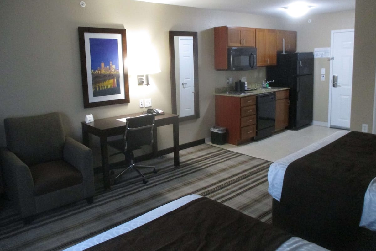 A hotel room with a bed, a desk, a chair, a kitchenette, and a framed picture on the wall.