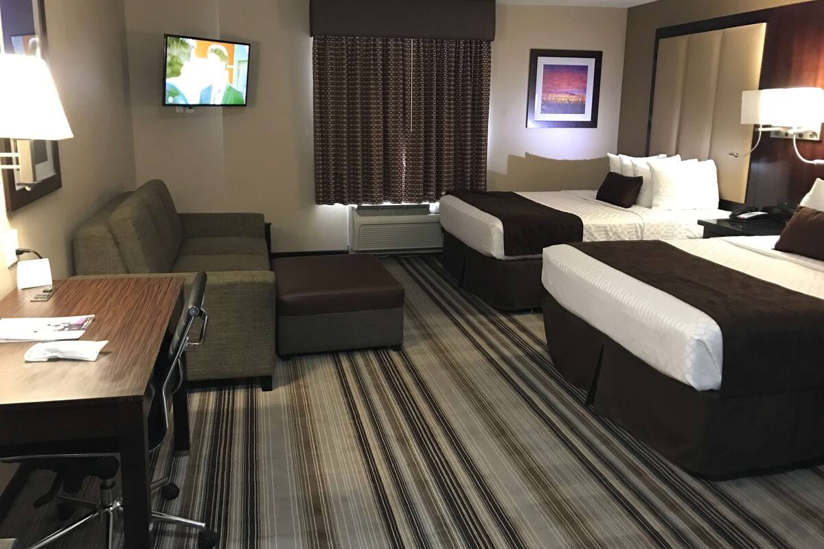 Ein Hotelzimmer mit einer Sitzecke, zwei Einzelbetten, einem Schreibtisch mit Stuhl, einem Fernseher, einem Fenster mit gemusterten Vorhängen und einem gerahmten Bild an der Wand.