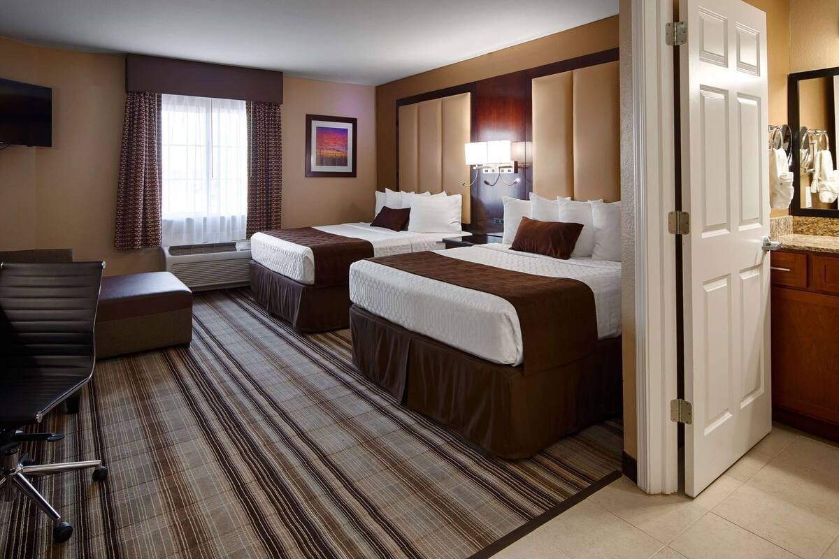 A hotel room with two beds, a desk, a chair, a TV, a window with curtains, a painting, and a bathroom visible through an open door.