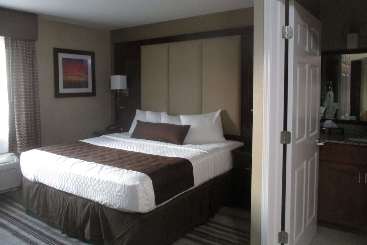 A hotel room with a large bed, a nightstand, a lamp, a framed picture on the wall, and a door leading to another room.