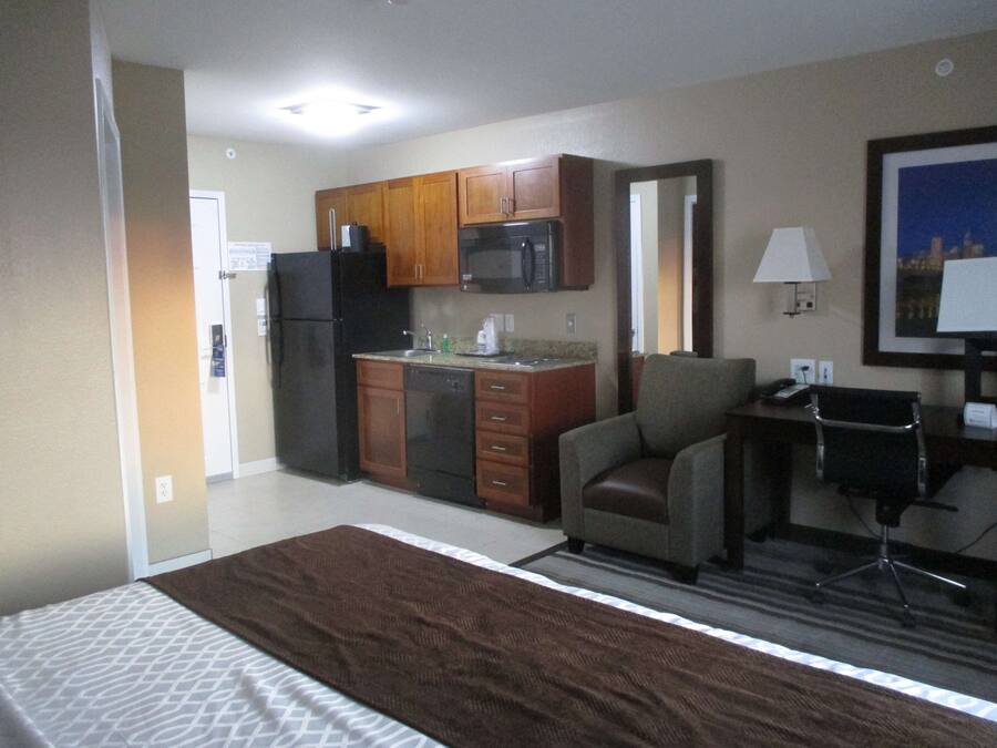 Una habitación de hotel con cama, kitchenette, una silla, escritorio y lámpara.