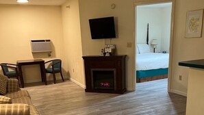A living room with a fireplace, a television, and a bed in the background. - Up North Hotel (formerly Voyager Inn) (St Ignace)