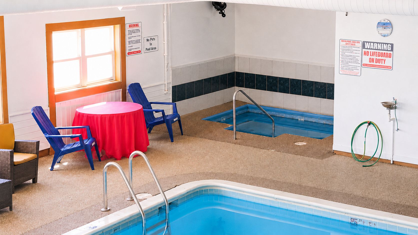 An indoor swimming pool area with a red table, blue chairs, and safety signs.