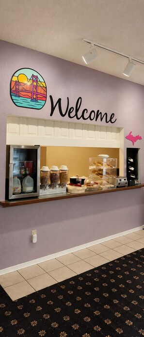 A reception area with a welcome sign, a refrigerator with beverages, a coffee station, and a wall-mounted light fixture.