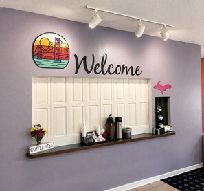 Zona de recepción con un cartel que dice 'Welcome', indicaciones de café y té, y una estantería con diversos artículos.