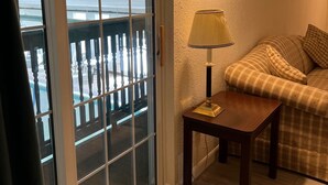 Una habitación con puerta corrediza de vidrio que da a un balcón, una mesita con lámpara, un sofá y una lámpara fijada a la pared.