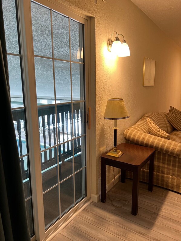 A room with a sliding glass door leading to a balcony, a small table with a lamp, a sofa, and a wall-mounted light fixture. - Up North Hotel (formerly Voyager Inn) (St Ignace)