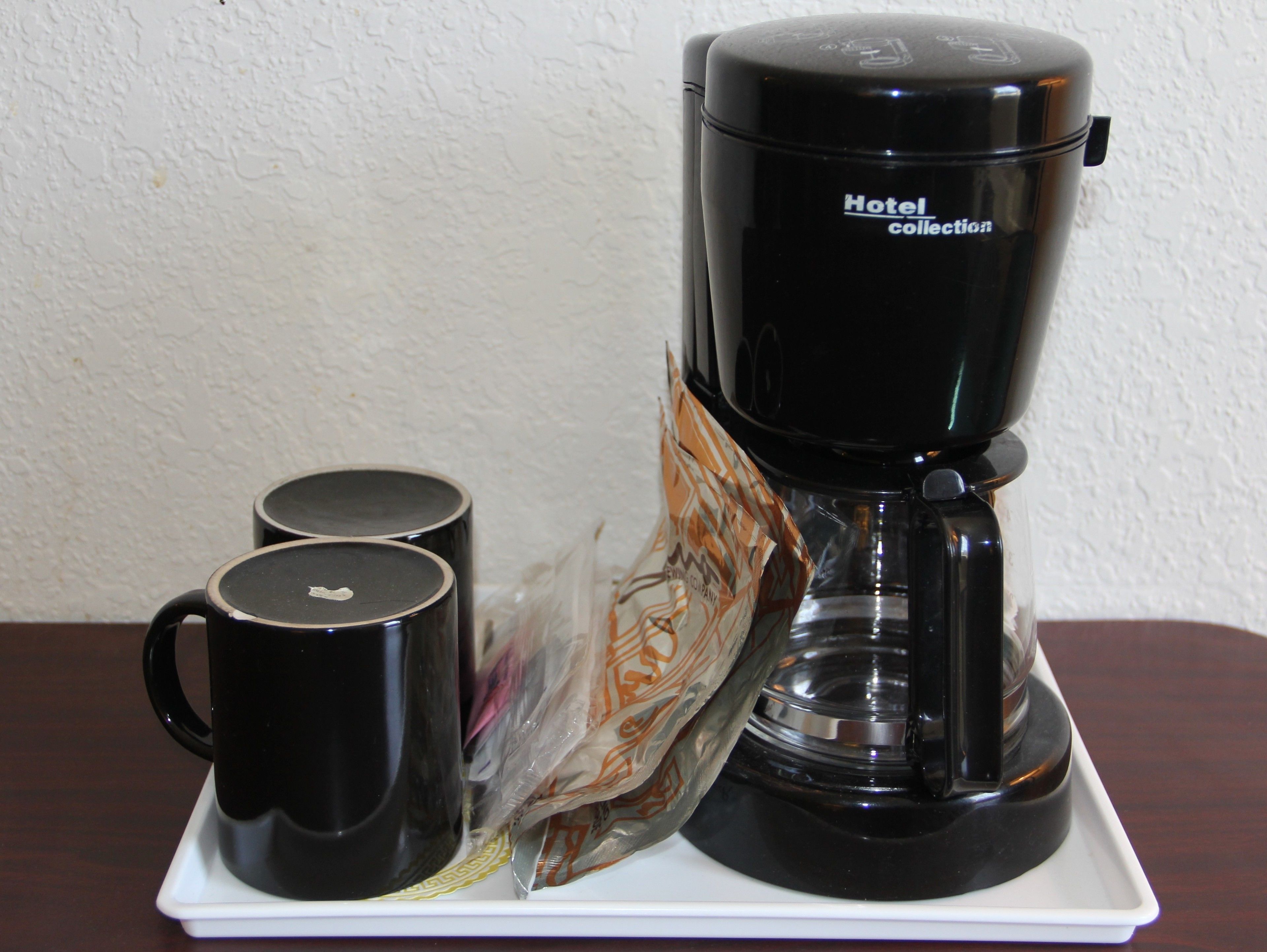 A black coffee maker labeled "Hotel Collection" on a white tray with two black mugs and a plastic bag containing a spoon.