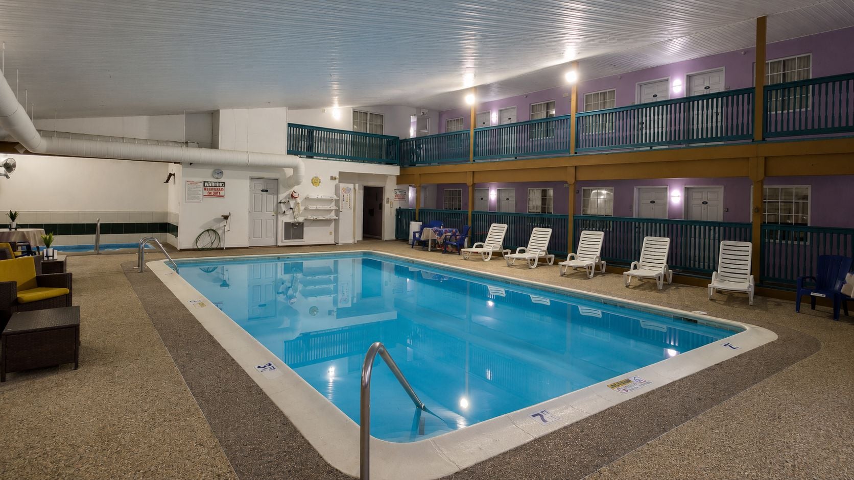 An indoor swimming pool with a blue water surface, surrounded by lounge chairs and a staircase leading to an upper level.