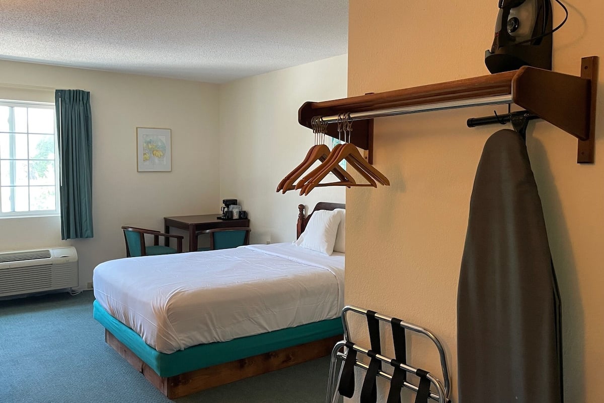 Ett hotellrum med en säng, ett skrivbord, en stol, en klädställning och en upphängd kappa.
