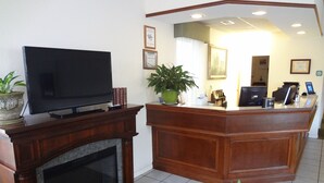 A hotel lobby with a TV, a reception desk, a fireplace, and a large potted plant. - Up North Hotel (formerly Voyager Inn) (St Ignace)