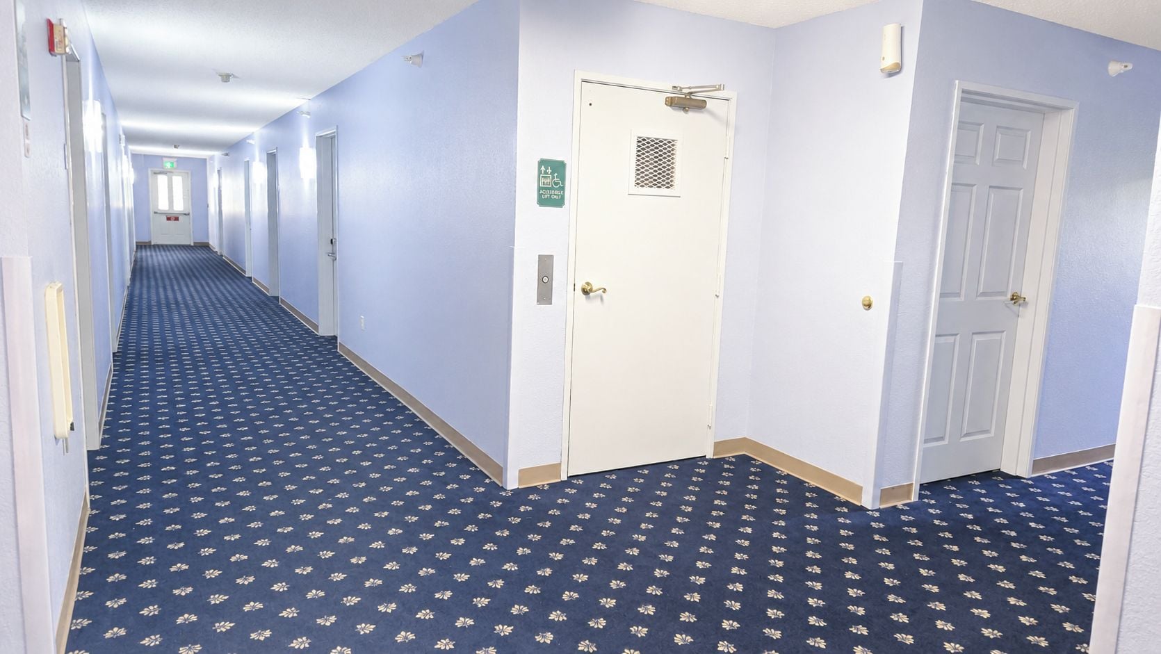 Un couloir d’hôtel avec un tapis à motifs, des portes blanches et un détecteur d’incendie au mur.
