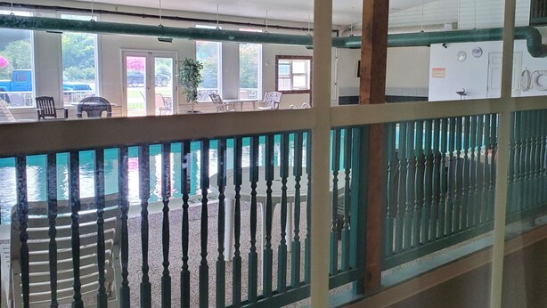 Vista de un área de piscina con muelle de madera y barandilla. Hay sillas y mesas alrededor de la piscina, y un edificio con ventanas al fondo.