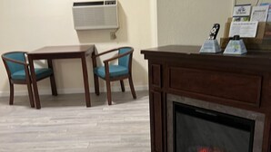 A living room with a fireplace, two wooden tables, two blue chairs, a mounted TV, and an air conditioning unit on the wall. - Up North Hotel (formerly Voyager Inn) (St Ignace)