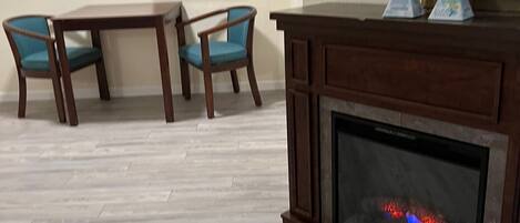 Una sala de estar con chimenea, dos mesas de madera, dos sillas azules, un televisor montado y un equipo de aire acondicionado en la pared.