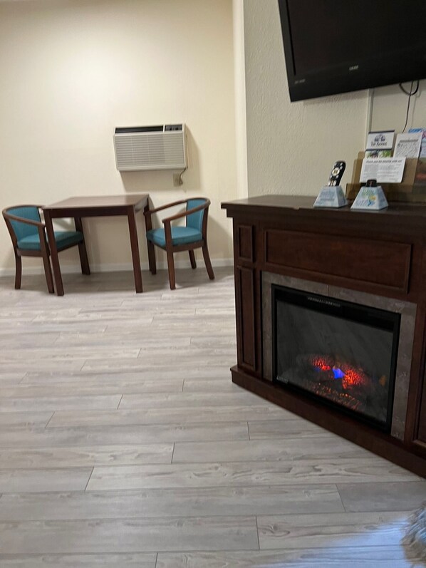 A living room with a fireplace, two wooden tables, two blue chairs, a mounted TV, and an air conditioning unit on the wall. - Up North Hotel (formerly Voyager Inn) (St Ignace)