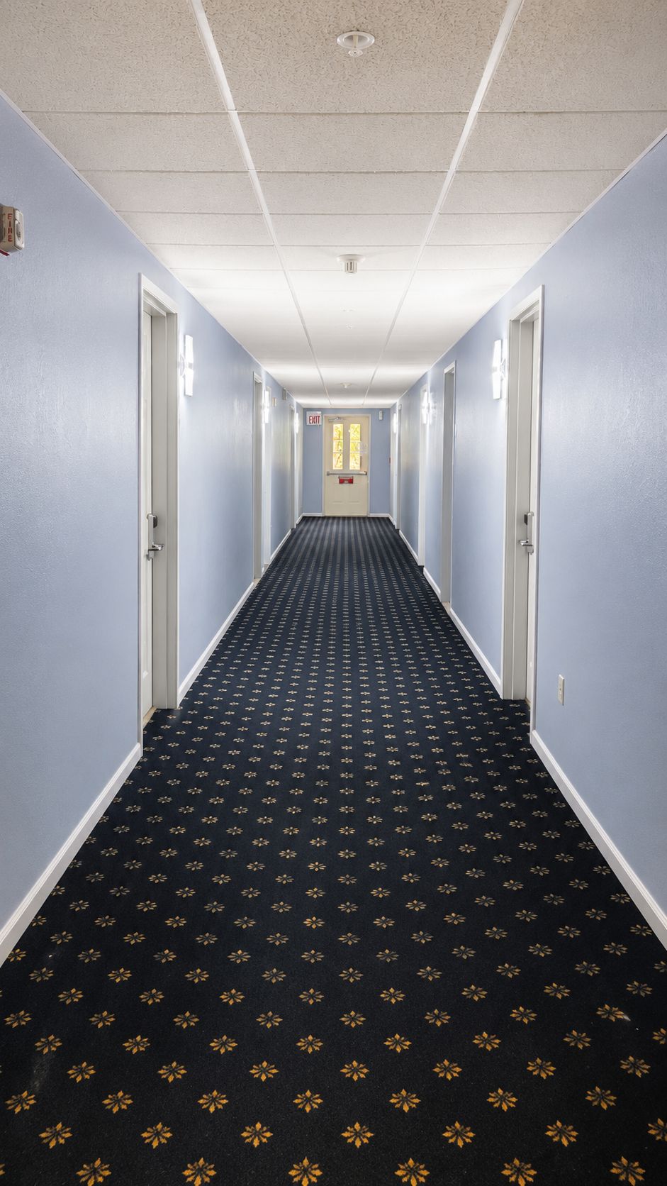 A hotel hallway with a patterned carpet, light blue walls, and doors leading to rooms.