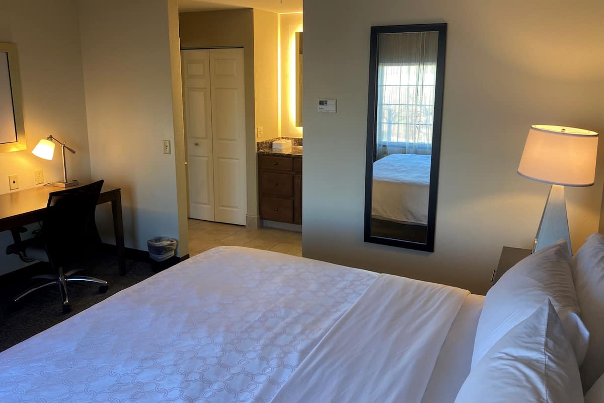 A hotel room with a bed, a desk with a chair, a mirror, and a lamp.