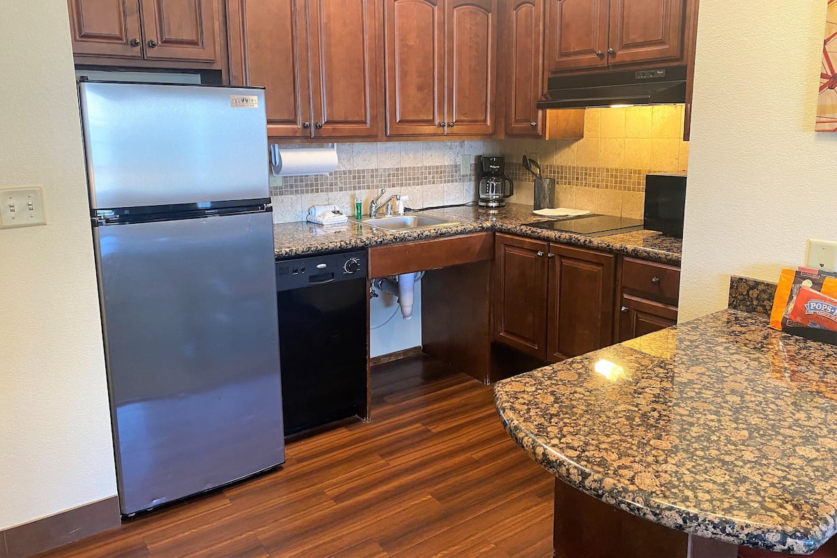 A kitchen with wooden cabinets, a stainless steel refrigerator, a dishwasher, granite countertops, and a coffee maker.