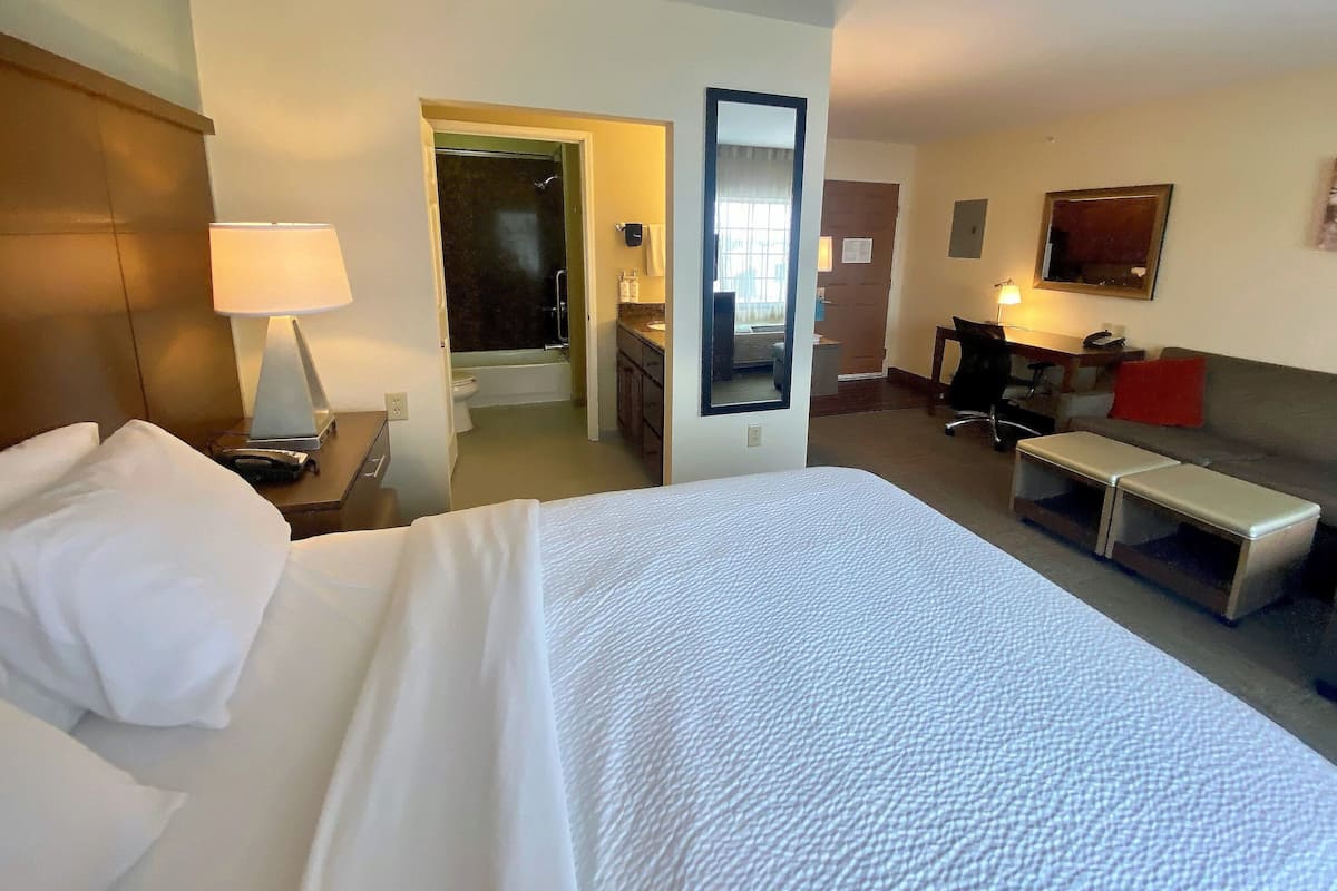 A hotel room with a large bed, a desk with a chair, a small table, a sofa, a mirror, and a bathroom visible through an open door.