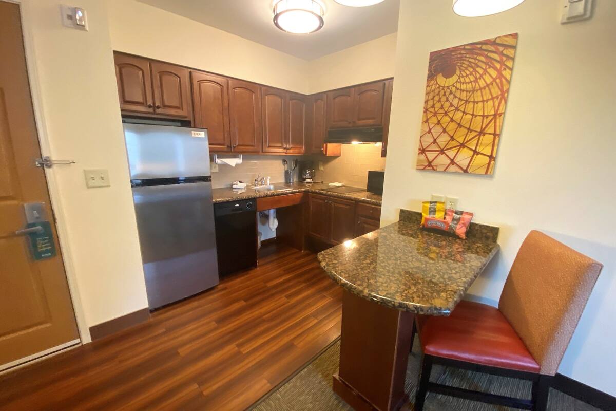 A compact kitchen with wooden cabinets, a stainless steel refrigerator, a dishwasher, and a granite countertop.