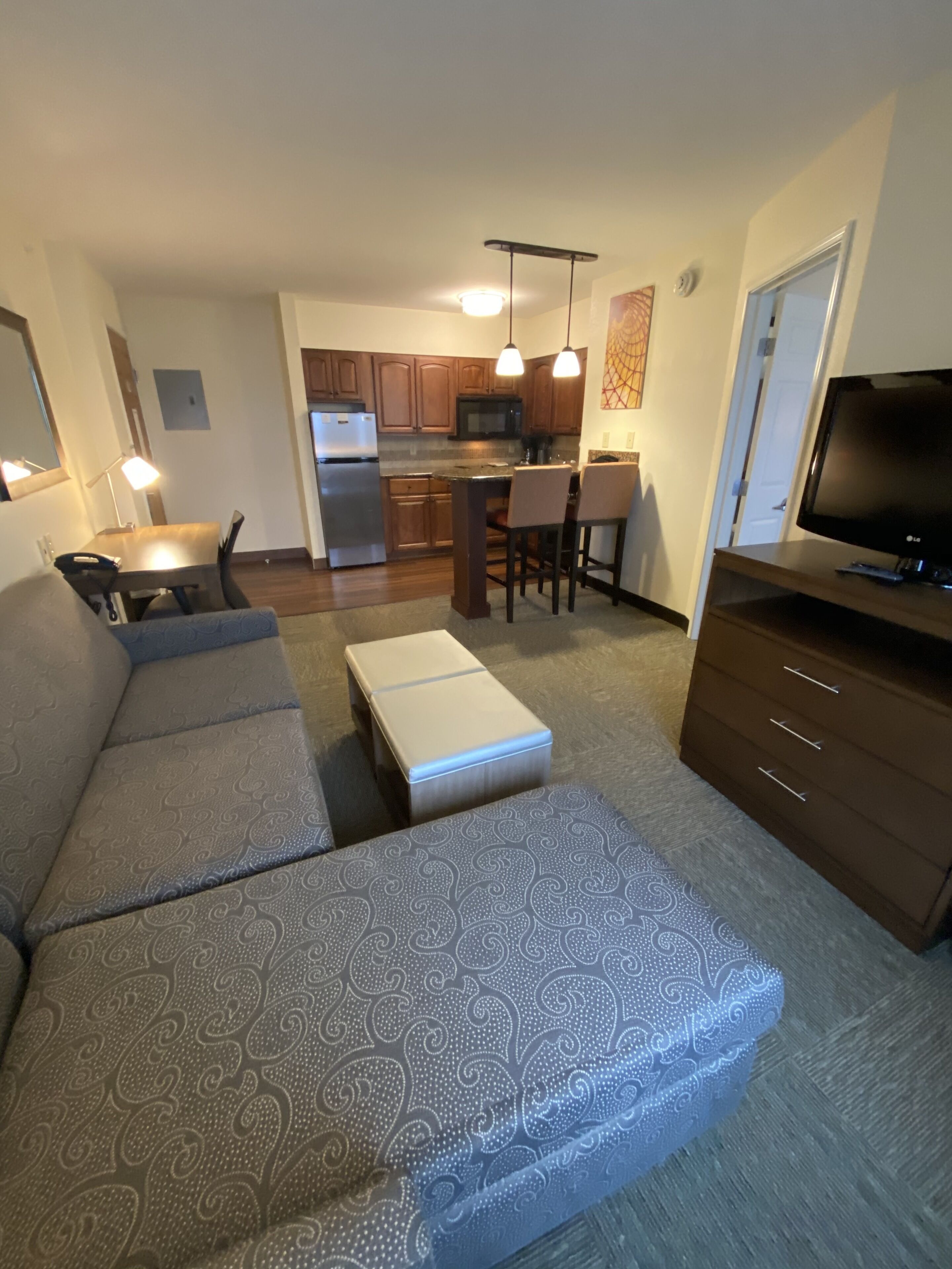 A living room with a sofa set, a television, and a kitchen area in the background.