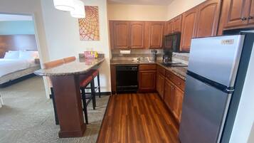 A compact hotel kitchen with wooden cabinets, a stainless steel refrigerator, and a small dining area with a bar stool.