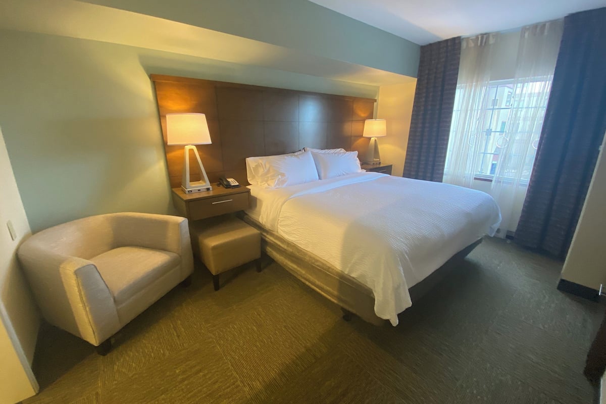 A hotel room with a large bed, two bedside tables with lamps, a chair, and a window with curtains.