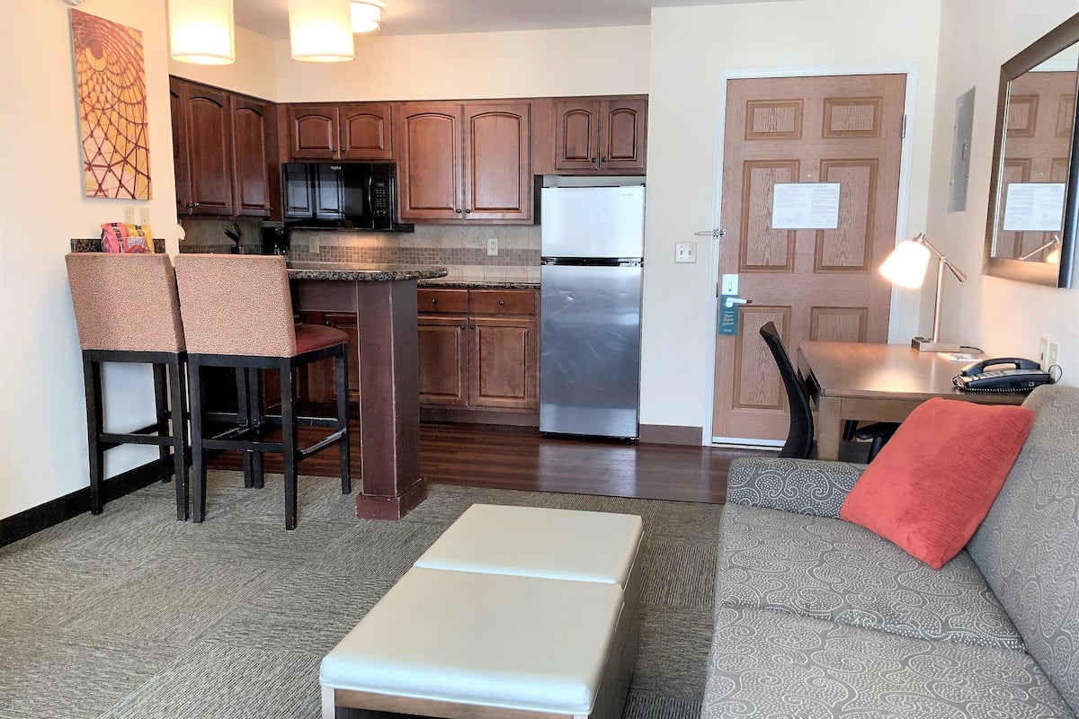 A compact kitchen with wooden cabinets, a refrigerator, and a microwave. There is a dining area with a table and chairs, and a sofa with a red pillow.