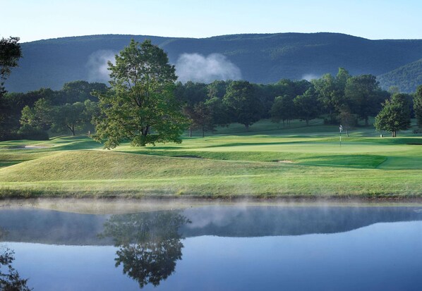 A serene golf course with a calm pond reflecting the landscape, surrounded by lush greenery and distant hills.