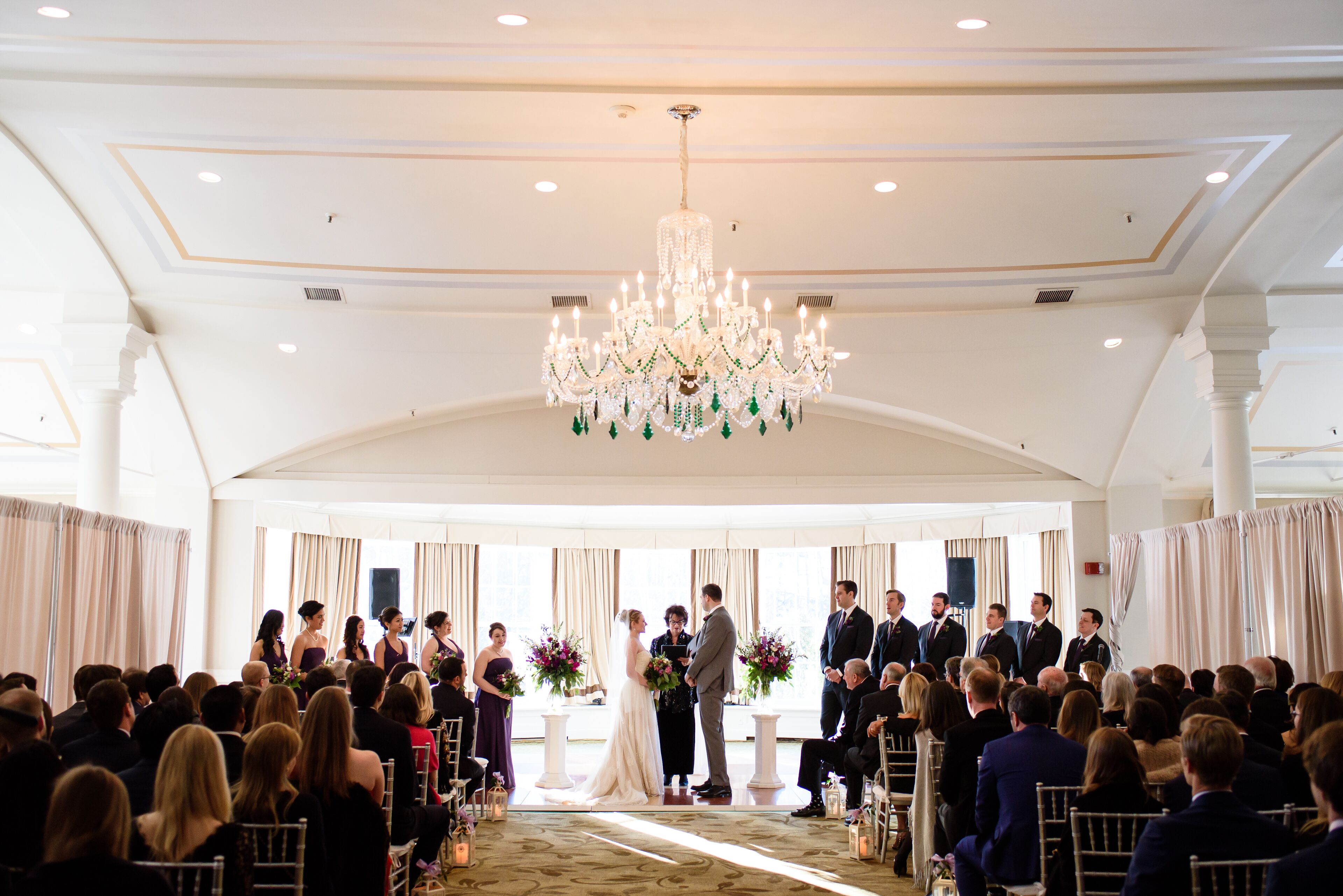 A wedding ceremony with guests seated on chairs arranged in rows, a couple standing at the altar, and a chandelier above.