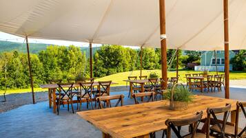 An outdoor dining area with wooden tables and chairs, a large canopy, and a view of a green landscape with trees and a house in the background.